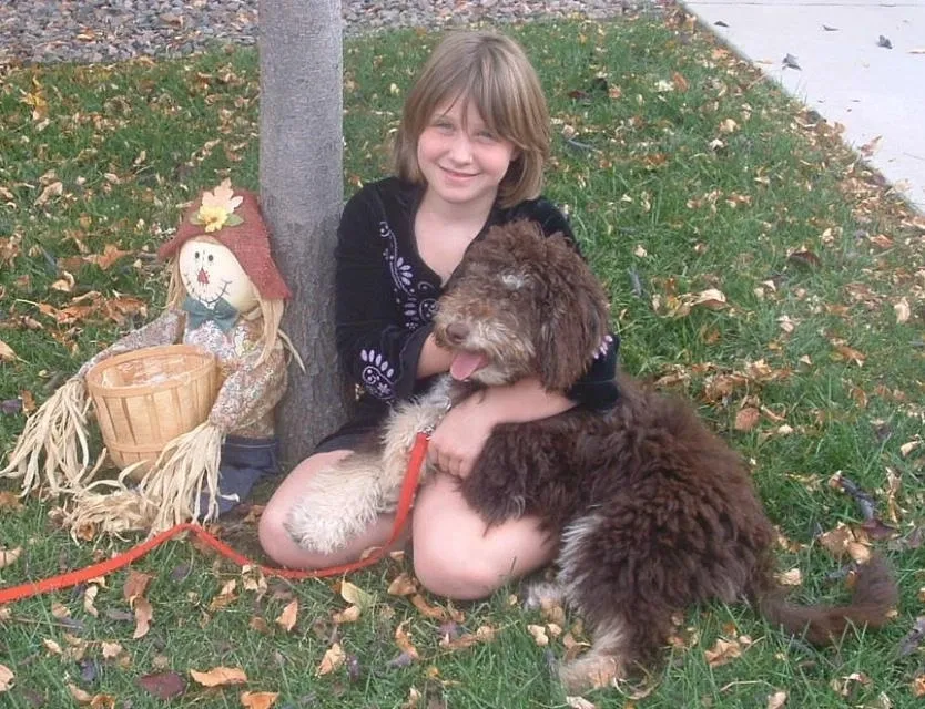 Young person sitting on a lawn hugging a brown dog next to a scarecrow decoration.